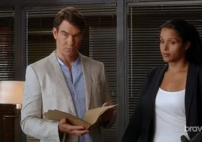 Homem de casaco claro e mulher de blazer em escritório. Ele olha sério, segurando documentos; ela observa atentamente. Clima profissional.