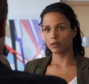 Mulher de cabelo encaracolado e blazer verde-oliva olha fixamente para a frente. Um homem está de costas para ela. Fundo de escritório com arte abstrata. Clima sério.