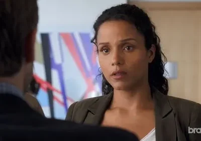 Mulher de cabelo encaracolado e blazer verde-oliva olha fixamente para a frente. Um homem está de costas para ela. Fundo de escritório com arte abstrata. Clima sério.