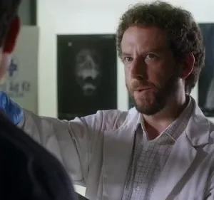 Cientista barbudo de jaleco branco fala com colega em laboratório. Raios-X de crânio e ossos ao fundo. Clima sério.