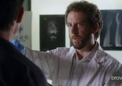 Cientista barbudo de jaleco branco fala com colega em laboratório. Raios-X de crânio e ossos ao fundo. Clima sério.