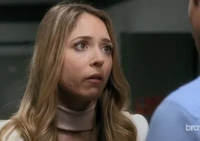 Jovem loira de olhos arregalados e blazer claro, olha preocupada para homem de costas. Clima tenso.