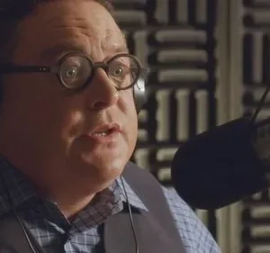 Homem de óculos e fones em estúdio, falando a um microfone. Parece surpreso ou engajado. Fundo com painéis acústicos.