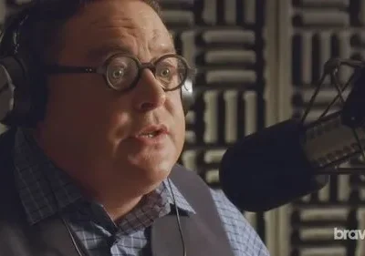 Homem de óculos e fones em estúdio, falando a um microfone. Parece surpreso ou engajado. Fundo com painéis acústicos.