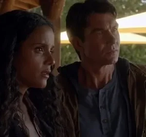 Mulher preocupada e homem focado em um deck. Ao fundo, guarda-chuvas amarelos, árvores e um lago sob luz solar.