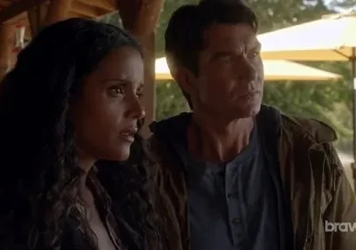 Mulher preocupada e homem focado em um deck. Ao fundo, guarda-chuvas amarelos, árvores e um lago sob luz solar.