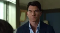Um homem de cabelo escuro, com blazer azul e camisa clara, olha com expressão séria. Fundo borrado mostra uma janela e outra pessoa.