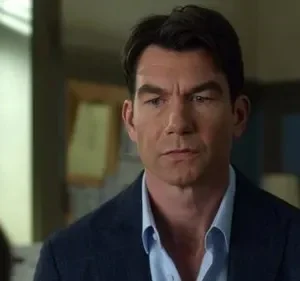 Um homem de cabelo escuro, com blazer azul e camisa clara, olha com expressão séria. Fundo borrado mostra uma janela e outra pessoa.