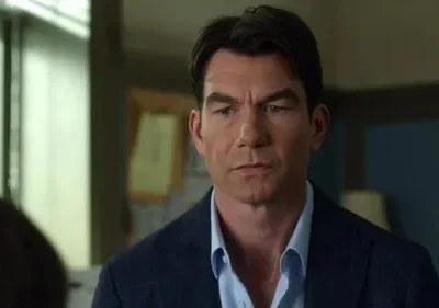 Um homem de cabelo escuro, com blazer azul e camisa clara, olha com expressão séria. Fundo borrado mostra uma janela e outra pessoa.