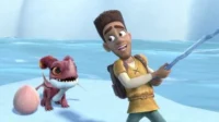 Homem animado de túnica amarela segura cajado de gelo, sorrindo. Ao lado, um dragão bebê vermelho e um ovo rosa, em paisagem nevada.