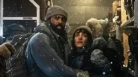 Duas pessoas em roupas de inverno, cobertas de neve, com expressões tensas dentro de um compartimento gelado. Clima de sobrevivência.
