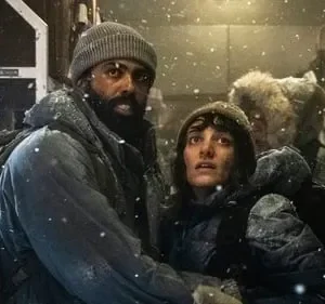 Duas pessoas em roupas de inverno, cobertas de neve, com expressões tensas dentro de um compartimento gelado. Clima de sobrevivência.