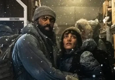 Duas pessoas em roupas de inverno, cobertas de neve, com expressões tensas dentro de um compartimento gelado. Clima de sobrevivência.