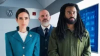 Três figuras sérias num ambiente tecnológico. Uma mulher de uniforme teal, um homem de dreads e barba em camisa verde, e um careca de terno.
