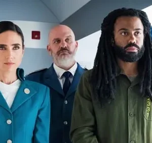 Três figuras sérias num ambiente tecnológico. Uma mulher de uniforme teal, um homem de dreads e barba em camisa verde, e um careca de terno.