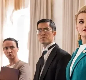 Quatro pessoas em ambiente formal. Uma mulher loira de jaqueta turquesa olha surpresa, os outros parecem sérios.