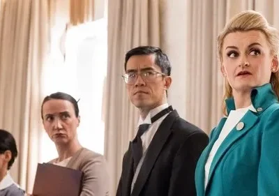 Quatro pessoas em ambiente formal. Uma mulher loira de jaqueta turquesa olha surpresa, os outros parecem sérios.