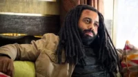 Homem com dreadlocks e barba, de pele escura, com ferimento na testa. Veste jaqueta cáqui e senta pensativo em ambiente interno.
