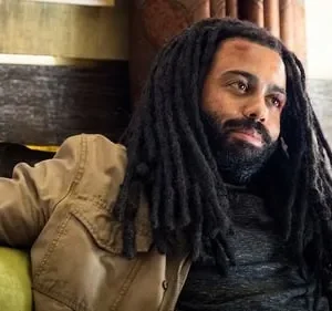 Homem com dreadlocks e barba, de pele escura, com ferimento na testa. Veste jaqueta cáqui e senta pensativo em ambiente interno.