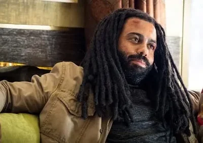 Homem com dreadlocks e barba, de pele escura, com ferimento na testa. Veste jaqueta cáqui e senta pensativo em ambiente interno.