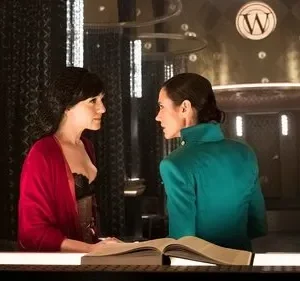 Duas mulheres em cenário luxuoso e escuro. Uma de vermelho, outra de verde-água, conversam seriamente sobre um livro.