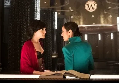 Duas mulheres em cenário luxuoso e escuro. Uma de vermelho, outra de verde-água, conversam seriamente sobre um livro.