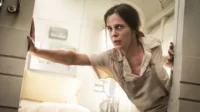 Mulher de uniforme bege com expressão de medo espreita de uma cabine branca e apertada. Clima de suspense.