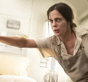 Mulher de uniforme bege com expressão de medo espreita de uma cabine branca e apertada. Clima de suspense.