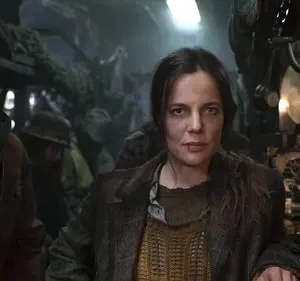 Homem de boné e mulher séria, com ferimento, em cenário sombrio e industrial/pós-apocalíptico. Clima de sobrevivência.