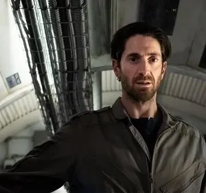 Homem barbudo de uniforme em nave espacial futurista. Ele tem expressão preocupada. Ambiente com cabos e luzes azuis.