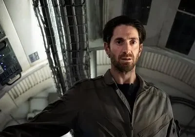 Homem barbudo de uniforme em nave espacial futurista. Ele tem expressão preocupada. Ambiente com cabos e luzes azuis.