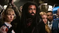 Homem com dreads e sangue no rosto, e mulher ensanguentada no meio de uma multidão tensa. Clima de urgência e perigo.