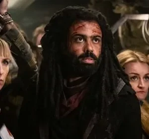 Homem com dreads e sangue no rosto, e mulher ensanguentada no meio de uma multidão tensa. Clima de urgência e perigo.