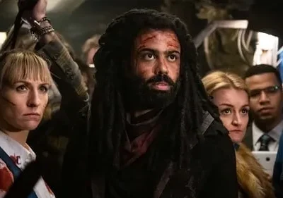 Homem com dreads e sangue no rosto, e mulher ensanguentada no meio de uma multidão tensa. Clima de urgência e perigo.