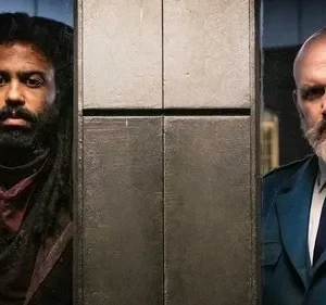 Dois homens sérios, um com dreads e roupa escura, outro careca com barba branca e uniforme militar, separados por um pilar metálico.