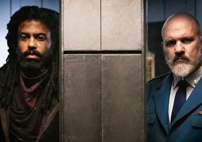 Dois homens sérios, um com dreads e roupa escura, outro careca com barba branca e uniforme militar, separados por um pilar metálico.