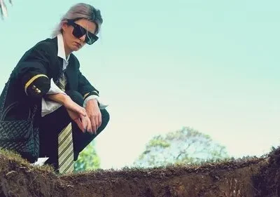 Pessoa de óculos escuros e blazer, agachada na beira de uma cova, olhando para baixo. O céu azul e árvores ao fundo criam um clima sombrio.
