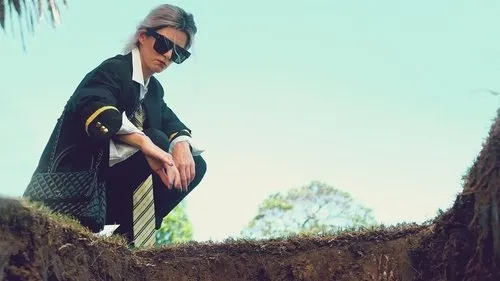 Pessoa de óculos escuros e blazer, agachada na beira de uma cova, olhando para baixo. O céu azul e árvores ao fundo criam um clima sombrio.
