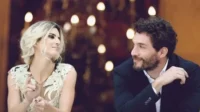 Homem barbudo e mulher loira trocam olhares e sorrisos. Ela usa blusa clara, ele terno escuro, com luzes quentes ao fundo.