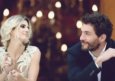 Homem barbudo e mulher loira trocam olhares e sorrisos. Ela usa blusa clara, ele terno escuro, com luzes quentes ao fundo.