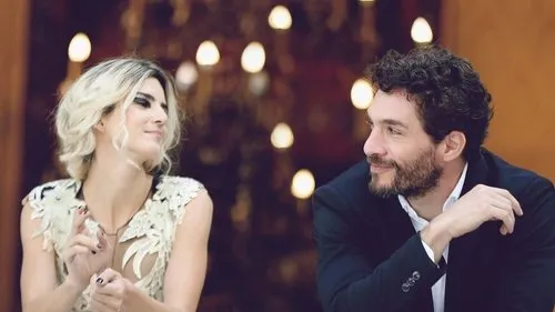 Homem barbudo e mulher loira trocam olhares e sorrisos. Ela usa blusa clara, ele terno escuro, com luzes quentes ao fundo.