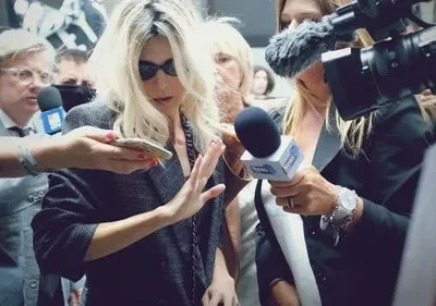 Mulher loira de óculos escuros e jaqueta cinza é cercada por jornalistas com microfones e câmeras, tentando afastar a atenção.