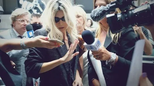 Mulher loira de óculos escuros e jaqueta cinza é cercada por jornalistas com microfones e câmeras, tentando afastar a atenção.