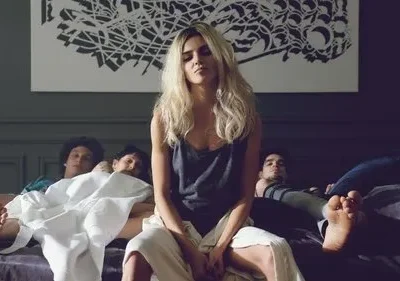 Mulher loira de expressão séria sentada na cama com três homens ao redor, parecendo dormir. Quarto escuro com arte abstrata na parede.