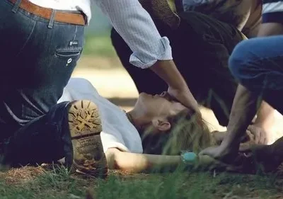 Uma pessoa deitada na grama é cercada por outras que se inclinam e tocam seu corpo, em um momento de socorro e drama.