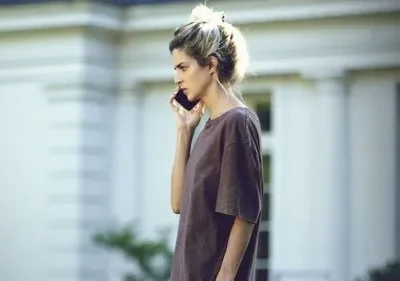 Uma mulher de cabelo claro e camiseta marrom fala ao telefone com semblante sério. Ao fundo, um prédio branco elegante.