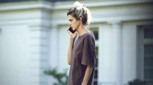 Uma mulher de cabelo claro e camiseta marrom fala ao telefone com semblante sério. Ao fundo, um prédio branco elegante.