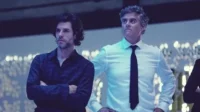 Dois homens com expressões sérias. Um de cabelo escuro, camisa escura; outro, grisalho, camisa branca e gravata. Fundo azul borrado com luzes.