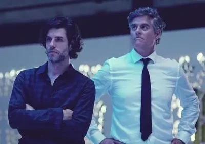 Dois homens com expressões sérias. Um de cabelo escuro, camisa escura; outro, grisalho, camisa branca e gravata. Fundo azul borrado com luzes.