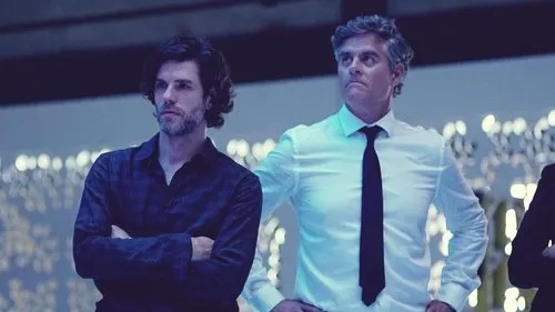 Dois homens com expressões sérias. Um de cabelo escuro, camisa escura; outro, grisalho, camisa branca e gravata. Fundo azul borrado com luzes.